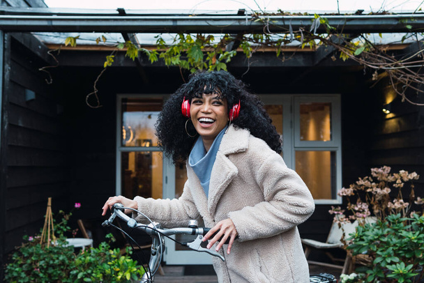 Widok z boku uśmiechnięta młoda Afroamerykanka z kręconymi ciemnymi włosami nosząca ciepły płaszcz spacerujący z rowerem podczas słuchania muzyki w słuchawkach w pobliżu domu jesienią - Zdjęcie, obraz
