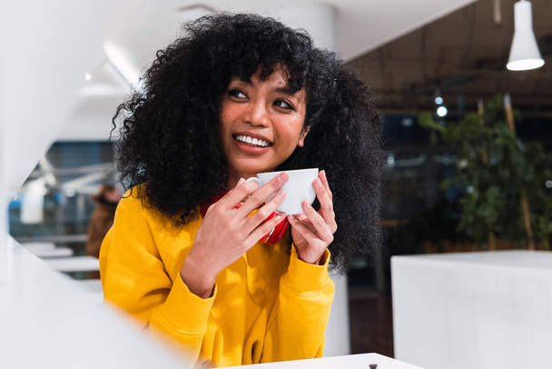 Contenida joven hembra étnica con el pelo largo Afro sonriendo y bebiendo taza de café en la cafetería ligera - Foto, imagen