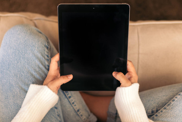Zbliżenie tabletu cyfrowego, który nierozpoznawalna kobieta trzyma w rękach, siedząc na kanapie w domu. - Zdjęcie, obraz