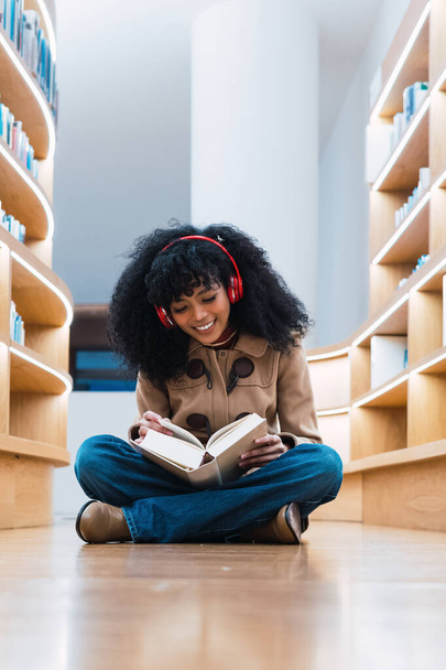 Happy African American female student in headphones reading book while sitting on floor in library and enjoying - Photo, Image