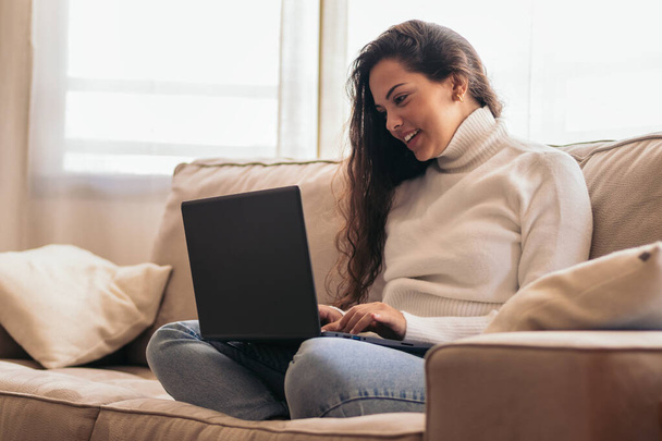 Evdeki kanepesinde oturan genç bir kadın, dizüstü bilgisayarıyla çalışıyor, yüzünde odaklanmış bir ifadeyle.. - Fotoğraf, Görsel