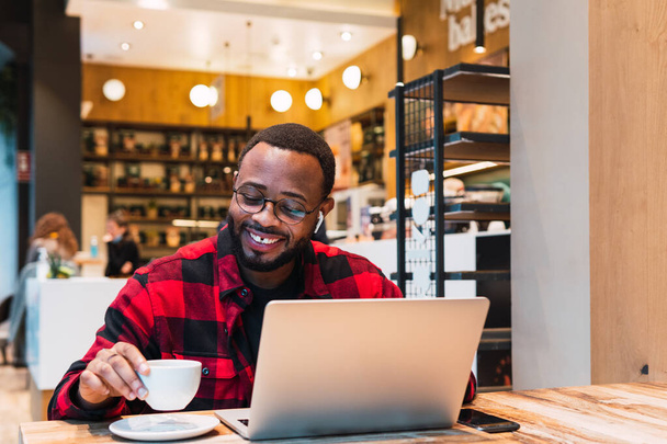 Afro-Amerikan sakallı erkek, kafede uzaktan çalışırken kulaklıkla netbook 'u tarıyor ve ekrana bakıyor. - Fotoğraf, Görsel