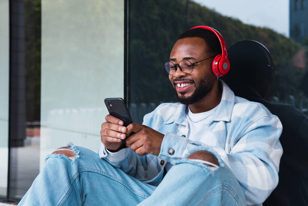 Szczęśliwy Afroamerykanin z telefonem komórkowym słuchający muzyki w słuchawkach bezprzewodowych siedząc przy ścianie nowoczesnego budynku - Zdjęcie, obraz