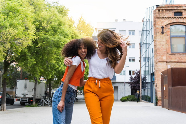 Веселые молодые многорасовые подруги в повседневной одежде обнимаются на городской тротуаре в летний день - Фото, изображение