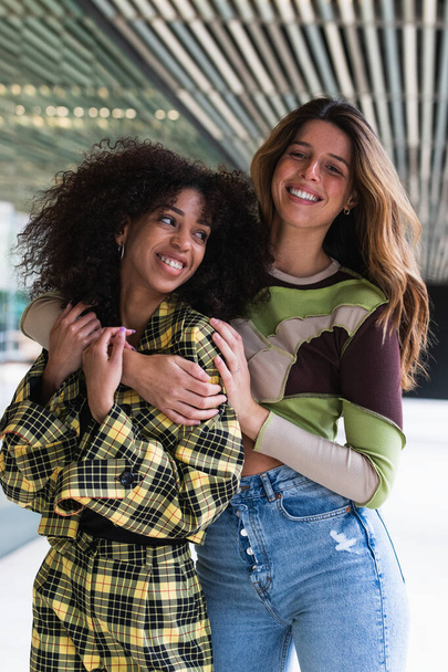 Jovem mulher sorridente abraçando alegre namorada étnica com penteado afro em terno xadrez na moda na cidade - Foto, Imagem