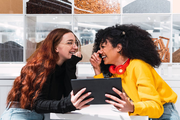 Oldalnézet vidám fiatal multiraciális női legjobb barátok alkalmi ruhák nevetve boldogan ül a modern kávézó táblagéppel a kezében - Fotó, kép