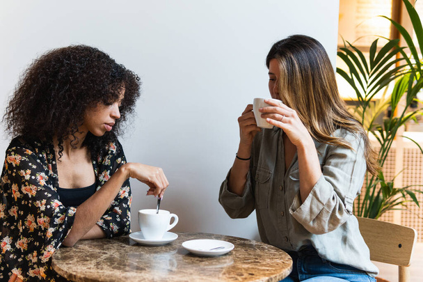 Fiatal multiracionális női barátok csésze forró italokkal, akik napközben az ebédlőasztalnál ülnek - Fotó, kép