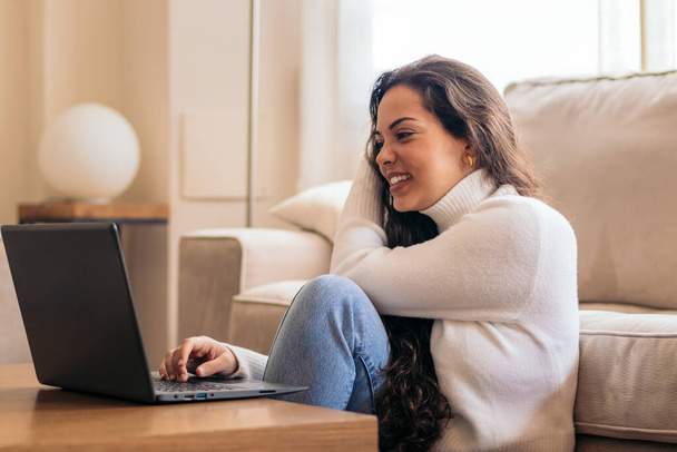Νεαρή γυναίκα στο σπίτι, εργάζεται στο φορητό υπολογιστή της με μια εστιασμένη έκφραση στο πρόσωπό της. - Φωτογραφία, εικόνα