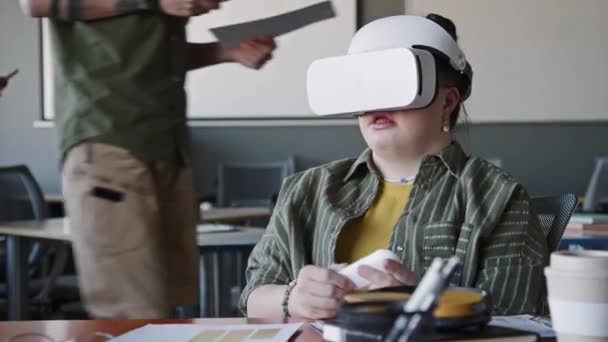 Девушка с синдромом Дауна сидит за столом в офисе и использует пульт дистанционного управления, исследуя дополненную реальность с помощью VR гарнитуры в течение рабочего дня - Кадры, видео