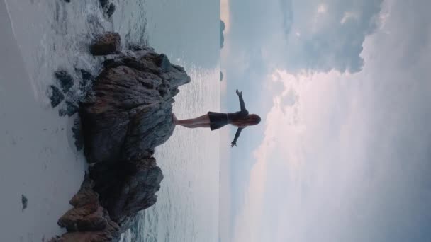 Вертикальное видео женщина стоит на прибрежном валуне возле океана, руки открыты морскому ветру. Сильный ветер от океана взъерошивает волосы и развевает платья. Женщина наслаждается нетронутыми и мощными элементами океана - Кадры, видео
