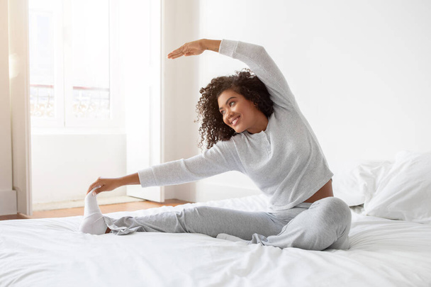 La mujer hispana está estirando su cuerpo mientras está acostada en una cama con sábanas blancas. Ella está extendiendo sus brazos y piernas para aflojar sus músculos después de despertar. - Foto, Imagen