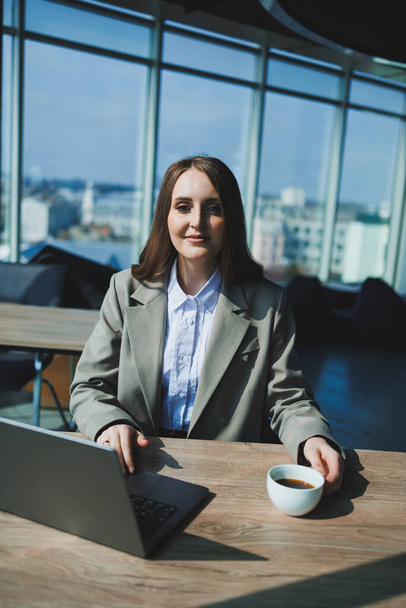 Mladá výkonná žena sedí u stolu s šálkem kávy a notebookem, zatímco pracuje v kanceláři. - Fotografie, Obrázek