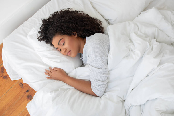 Mulher hispânica está deitada em uma cama com um edredão branco cobrindo-a, aparecendo relaxado e confortável em um ambiente de quarto, vista acima - Foto, Imagem