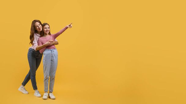 Mother and daughter dressed in casual wear, stand closely together against a vibrant yellow backdrop, pointing at copy space while they both smile brightly, sharing a lighthearted moment. - Photo, Image