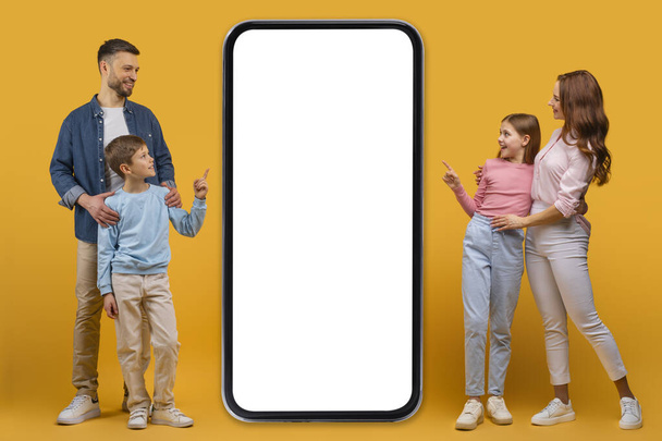 Hymyilevä isä, äiti, tytär ja poika seisovat yhdessä ylisuuren älypuhelimen vieressä, jossa on tyhjä näyttö, mahdollisesti vuorovaikutuksessa digitaalisen sovelluksen tai mainoksen kanssa - Valokuva, kuva