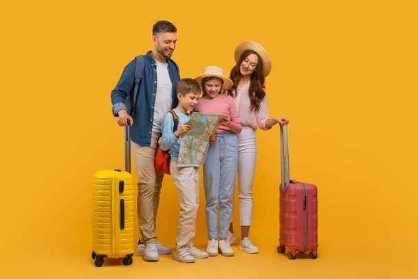 Весела сім'я, з двома дорослими та двома дітьми, готується до поїздки. Батьки стоять з валізами, а діти кружляють навколо карти, жовтий студійний фон - Фото, зображення