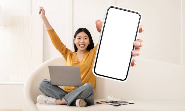 Una giovane donna asiatica allegra, vestita con un top giallo casual e jeans, si siede a gambe incrociate su un accogliente divano bianco con il suo computer portatile, presentando con entusiasmo uno smartphone con uno schermo bianco - Foto, immagini