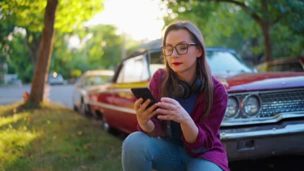 Mujer con auriculares se agacha abajo junto al coche rojo vintage y el uso de teléfono inteligente en la calle verde en el día de verano. La tendencia a la moda.  - Metraje, vídeo