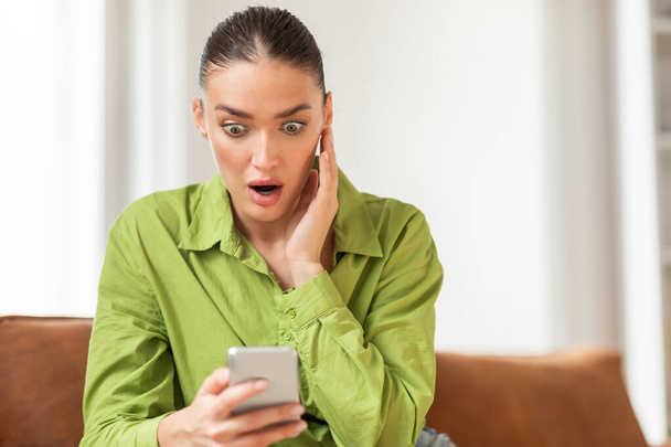 Eine junge Frau in einer auffallend grünen Bluse sitzt drinnen und blickt überrascht und ungläubig auf ihr Smartphone. Ihre Augen sind weit aufgerissen, und ihre Hand wird an ihre Wange gedrückt. - Foto, Bild