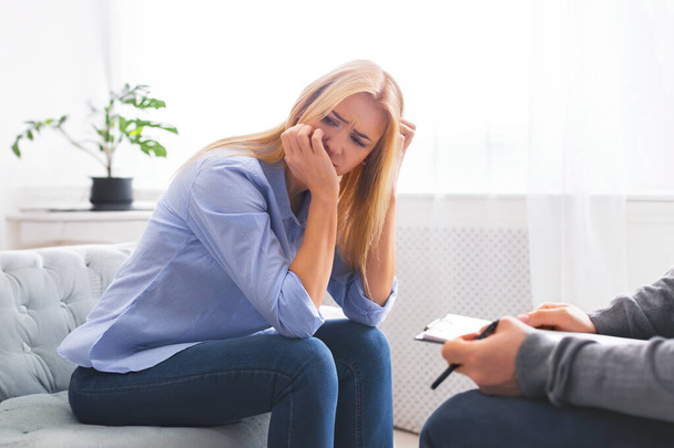 Una mujer se sienta en el borde de un sofá, su mano cubriéndose la cara, posiblemente secándose las lágrimas, expresando angustia o frustración durante una sesión de asesoramiento - Foto, Imagen