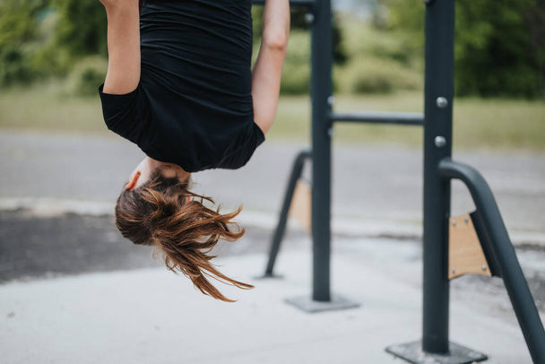 Динамичная сцена, где женщина занимается упражнениями вверх тормашками на фитнес-станции парка, демонстрируя силу и гибкость. - Фото, изображение