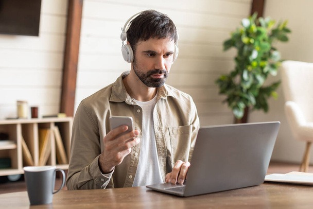 Ein Mann sitzt an einem Holztisch und tippt aufmerksam mit seinem Laptop, während er Kopfhörer trägt. Gleichzeitig hält er ein Smartphone in der rechten Hand - Foto, Bild