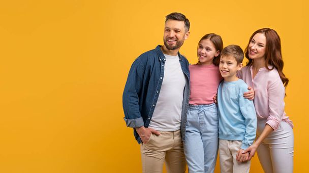 Šťastná rodina se dvěma rodiči a dvěma dětmi se objímají na zářivě žlutém pozadí. Vypadají vesele a spokojeně, dívají se na kopírovací prostor - Fotografie, Obrázek
