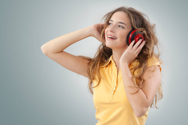 Ritratto di giovane donna che ascolta musica attraverso cuffie rosse, guarda a lato con gli occhi azzurri, mentre è isolata su uno sfondo grigio con spazio per copiare - Foto, immagini