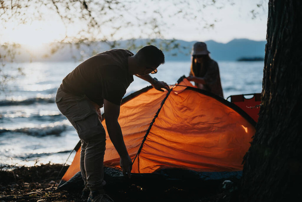 Twee vrienden genieten van hun weekend buiten als ze een campingtent opzetten bij een prachtig meer bij zonsondergang, omarmen ontspanning en saamhorigheid. - Foto, afbeelding