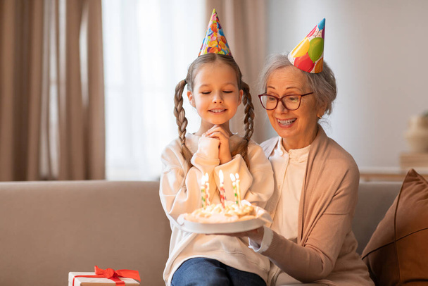 Літня жінка і молода дівчина сидять на дивані, перед ними торт на день народження. Жінка запалює свічки на торт, поки дівчина охоче дивиться - Фото, зображення