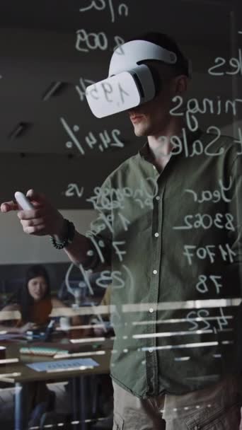 Вертикальний знімок молодої людини в гарнітурі VR, що стоїть за скляною стіною в офісі і використовує пульт дистанційного керування при розробці додатка доповнення реальності в IT-компанії - Кадри, відео