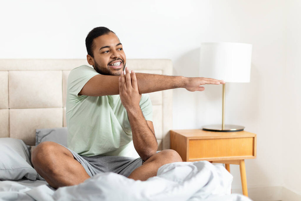 Молодой афроамериканец сидит на кровати в повседневной одежде и растягивается с довольной улыбкой - Фото, изображение