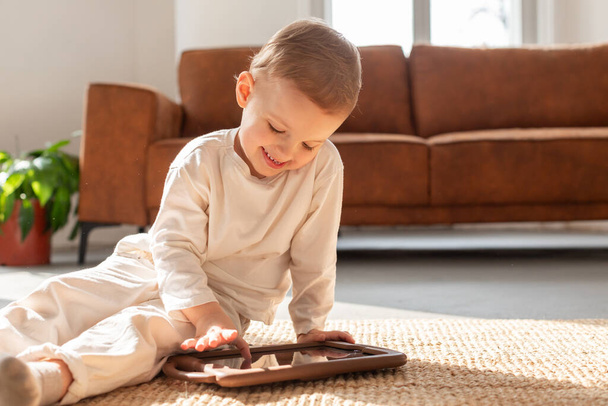Młody chłopiec siedzi na podłodze pochłonięty grą na tablecie. Skupia się na ekranie, stukając i przesuwając z koncentracją - Zdjęcie, obraz