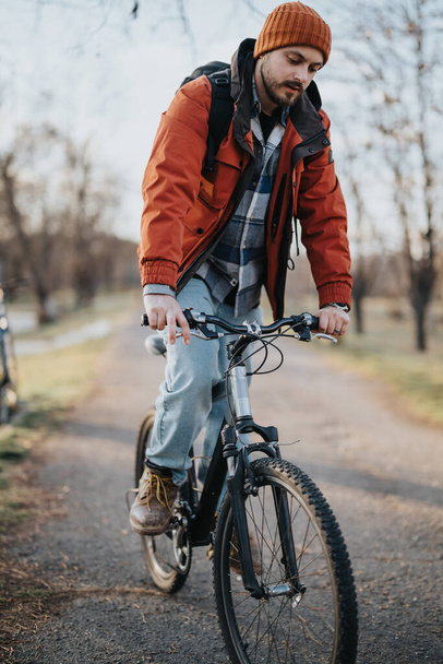 Een jonge, nonchalant geklede man vindt plezier tijdens het fietsen op een pad in een serene parkomgeving. - Foto, afbeelding