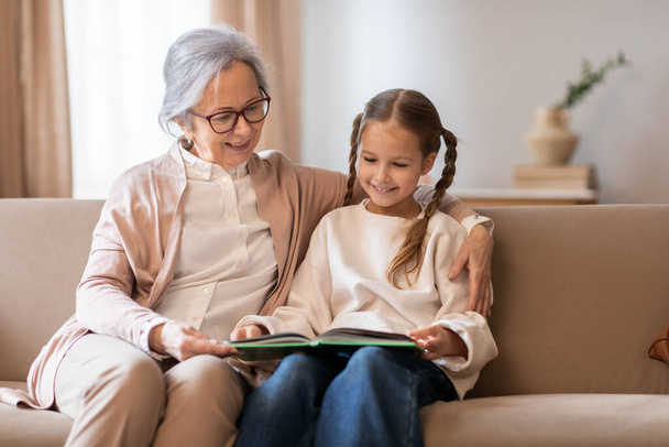 Eine ältere Frau und ein junges Mädchen sitzen zusammen auf einer Couch und sind in ein Buch vertieft, das sie gemeinsam lesen. Die Hand der Großmutter ruht auf dem Buch und führt das junge Mädchen - Foto, Bild