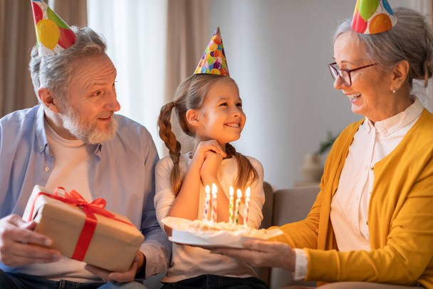 Syntymäpäivähattuun pukeutunut nuori tyttö hymyilee kirkkaasti, kun hänen isovanhempansa, jotka myös pukeutuvat juhlahattuihin, esittelevät hänelle syntymäpäiväkakun, joka on koristeltu valaistuilla kynttilöillä. - Valokuva, kuva
