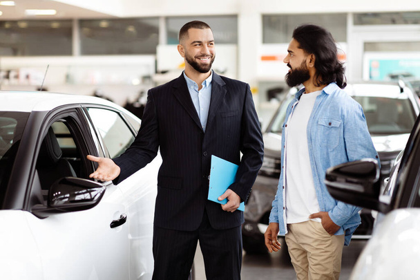 Un vendeur de voitures habillé intelligemment, avec un comportement amical, se tient à côté d'une nouvelle voiture brillante dans un showroom de véhicules bien éclairés, s'engageant avec un client potentiel indien qui tient une brochure. - Photo, image
