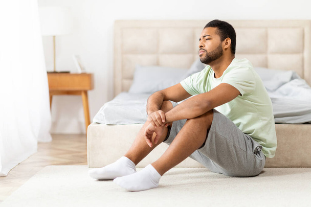 Ένας νεαρός Αφροαμερικάνος κάθεται στο πάτωμα δίπλα στο κρεβάτι σε μια ηλιόλουστη κρεβατοκάμαρα, φαίνεται αναστατωμένος, απογοητευμένος, καταθλιπτικός, αντίγραφο χώρου - Φωτογραφία, εικόνα