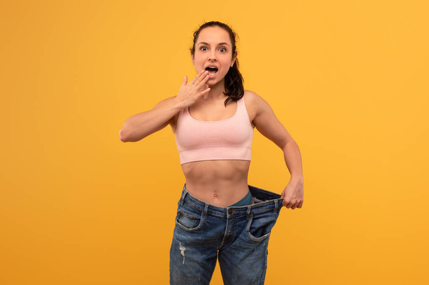 Молода жінка стоїть на тлі яскраво-жовтого фону, виражаючи сюрприз і досягнення, оскільки вона тримає пару великих джинсів, що свідчить про значну втрату ваги. - Фото, зображення