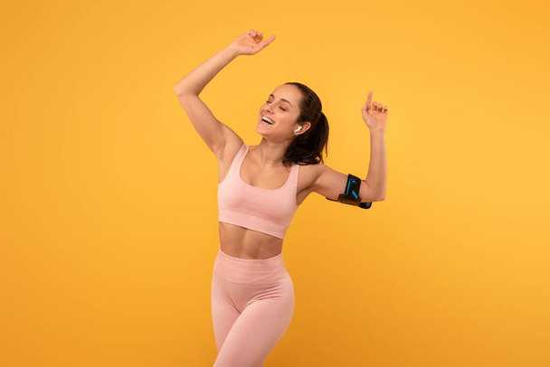 Μια νεαρή γυναίκα με ένα ακτινοβόλο χαμόγελο συλλαμβάνεται στη μέση του χορού σε ένα φωτεινό κίτρινο φόντο, ντυμένος με φόρμα-τοποθέτηση αθλητικών ενδυμάτων, και είναι σαφώς απολαμβάνοντας σε μια στιγμή θριάμβου ή εορτασμού - Φωτογραφία, εικόνα
