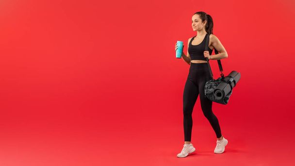 A fit woman dressed in black sportswear confidently strides forward while holding a yoga mat and gym bag. Her white sneakers contrast with the bright red backdrop - Photo, Image