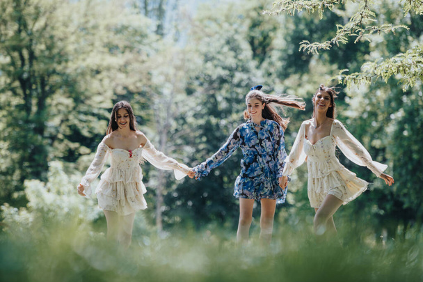 Onnelliset nuoret naiset tyylikkäissä asuissa juoksevat iloisesti ja pitävät kädestä auringonvalaistussa puistossa, jossa vallitsee vapaus ja ystävyys. - Valokuva, kuva