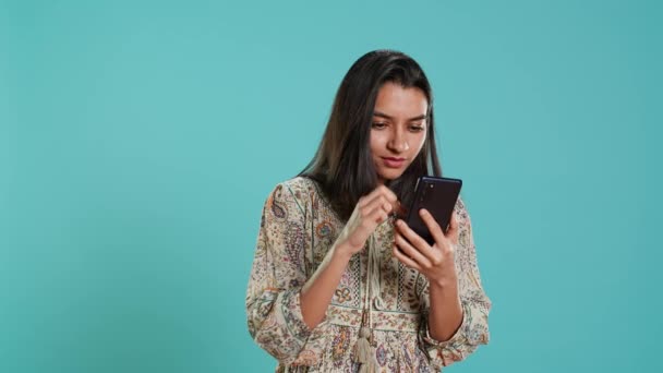 Woman happily scrolling on phone touchscreen to check social media feed, amazed by photo. Indian person browsing internet websites, astonished by content, studio background, camera A - Footage, Video