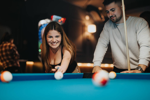 Eine glückliche junge Frau spielt Pool, während ein Mann zusieht, beide genießen einen lustigen Abend zusammen in einer Bar mit sanftem Licht gefüllt. - Foto, Bild