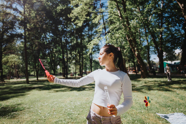 zorgeloze jonge volwassen vrouw spelen met een bubble wand buiten in een zonnig park setting, belichamen vrije tijd en geluk. - Foto, afbeelding