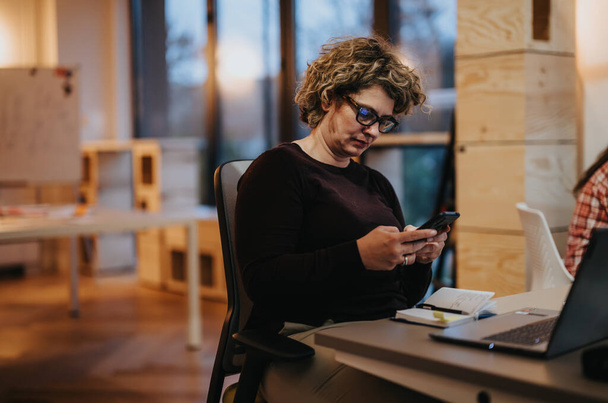 Una mujer de negocios madura se concentra en su teléfono durante un descanso en un espacio de oficina colaborativo con colegas de todo. - Foto, imagen