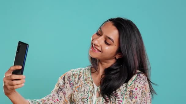 Jolly intialainen nainen hymyilee selfietä, lähettämällä ne sosiaalisessa mediassa matkapuhelimella. Iloinen tyttö ottaa kuvia puhelimella selfie kamera, eristetty studio tausta, kamera B - Materiaali, video