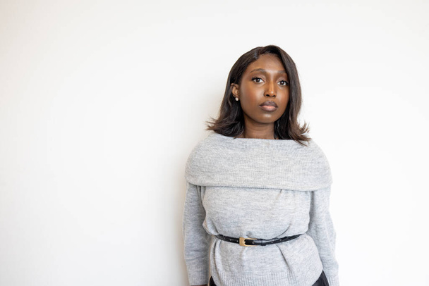This image features a young Black woman standing against a plain white background, dressed in a chic grey sweater and belted at the waist. Her expression is serene and thoughtful, conveying a sense of - Photo, Image