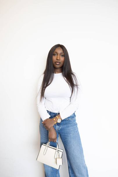 This portrait features a young Black woman standing against a plain white background, exuding elegance and confidence. She is dressed in a classic white long-sleeve top and casual blue jeans - Photo, Image