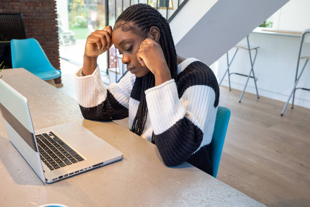 Молодая чернокожая женщина выглядит напряженной во время работы над ноутбуком в современном офисном пространстве. Она держит голову обеими руками, демонстрируя момент разочарования или концентрации. Офис имеет - Фото, изображение